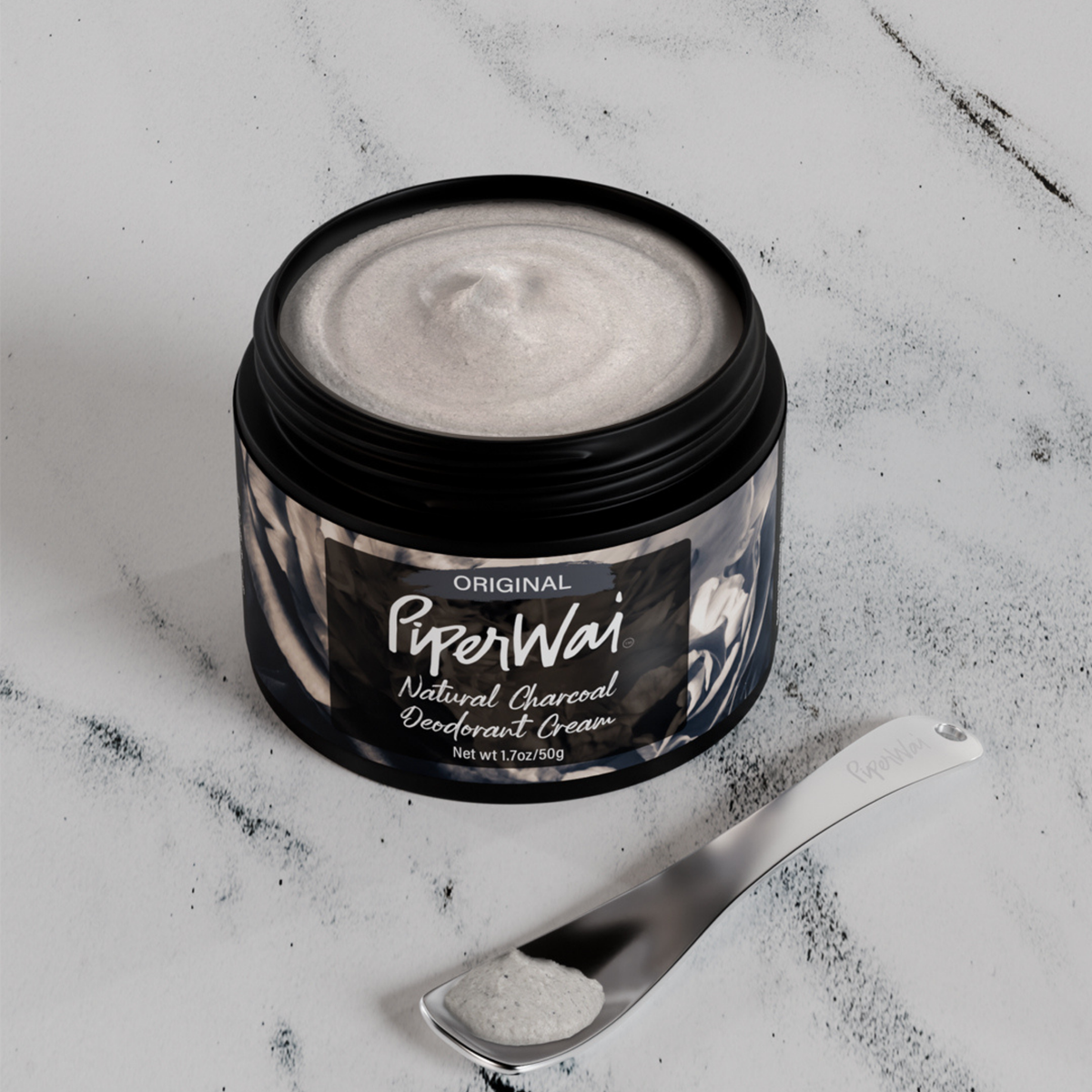 Metal cosmetic spoon with 'PiperWai' branding with an open jar of PiperWai deodorant cream against a textured background. 