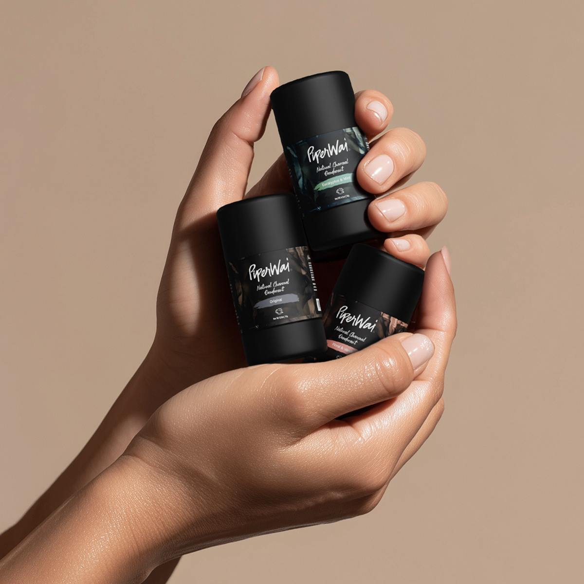 Hand holding three black mini deodorant sticks with visible branding against a beige background. 