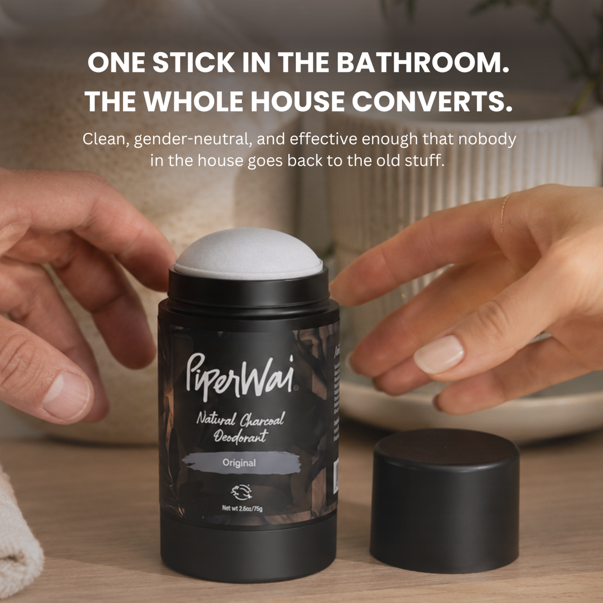 A black cylindrical deodorant stick container labeled PiperWai Natural Charcoal Deodorant, shown with a white cap on top and a separate black cap beside it on a tabletop. Two hands are reaching toward the product.