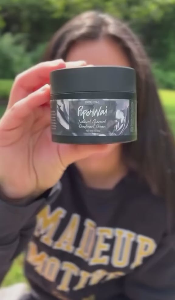 Woman outdoors in a dark sweatshirt applying PiperWai cream deodorant, then blowing a kiss to the camera